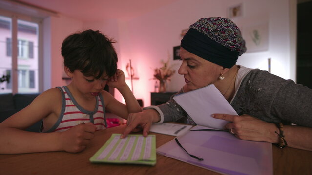 Mother undergoing chemotherapy helping child with homework at home, headscarf parent guiding learning at table, homeschooling moment showing care, patience and resilience