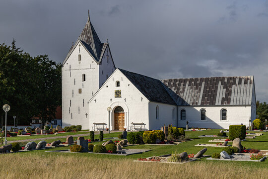 View of Romo Church featuring its white facade and pointed tower surrounded by a cemetery with gravestones under a cloudy sky in Romo, Denmark.