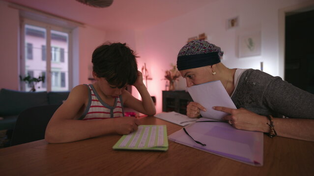 Mother undergoing chemotherapy helping child with homework at home, headscarf parent guiding learning at table, homeschooling moment showing care, patience and resilience