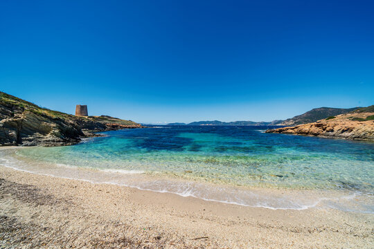 View of Spiaggia di Piscinni with turquoise water and the historic Torre di Piscinni tower on the rocky cliffs under a clear blue sky in Domus De Maria, Sardegna, Italy.