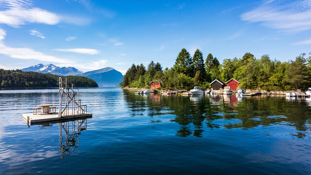View of a floating wooden platform on a calm fjord with traditional red boathouses and snow-capped mountains in Alesund, More og Romsdal, Norway.