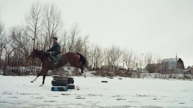 White rider galloping across snowy field on makeshift course, equestrian student navigating tire markers, steady canter with hooves kicking snow, rural skyline and bare trees, focused posture