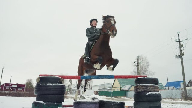 Snowy rural horse training for rider education, single rider clears makeshift fence on chestnut mount, village roofs and poles in background, overcast light highlights posture, technique,
