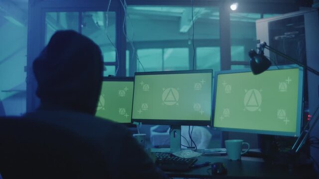 Man in the hood and the cap is sitting and working on stealing data at the computer as haker. Hideout Place has Dark Atmosphere.