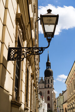 View of an ornate vintage street lamp mounted on a historic building facade leading towards the St. Nicholas Basilica tower under a blue sky in Trnava, Trnava Region, Slovakia.