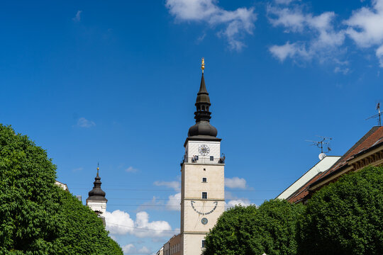 View of the historic City Tower with its distinctive dark spire and golden statue framed by lush green trees under a blue sky in Trnava, Trnava Region, Slovakia.