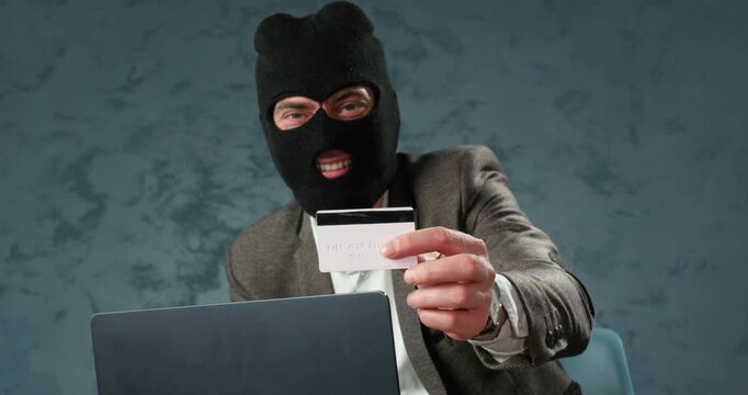 Scammer in balaclava laughs while showing bank card over laptop during fraud scheme. Happy man presents payment card mocking victim during online card deception
