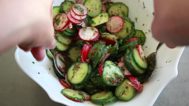 Close-up of preparing a simple summer salad by tossing sliced cucumbers and radishes with mustard dressing in a large salad bowl