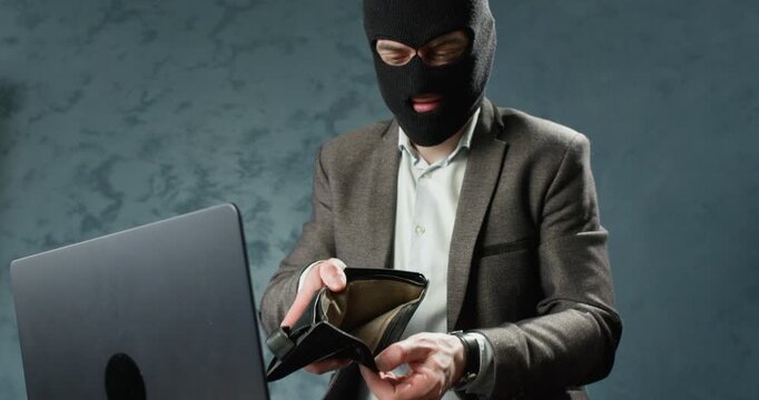 Masked online scammer opens empty leatyher wallet while sitting near laptop during online scam. Man shows lack of money using gesture to mock victim financial loss