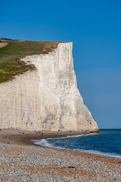 View of Seven Sisters Cliffs with towering white chalk cliffs, a pebble beach with people, and the calm blue sea under a clear sky in Exceat, England, United Kingdom.