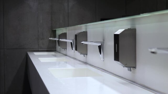 Minimalist interior of a modern public bathroom featuring a row of white ceramic sinks, chrome automatic faucets, and liquid soap dispensers mounted on a pristine white wall