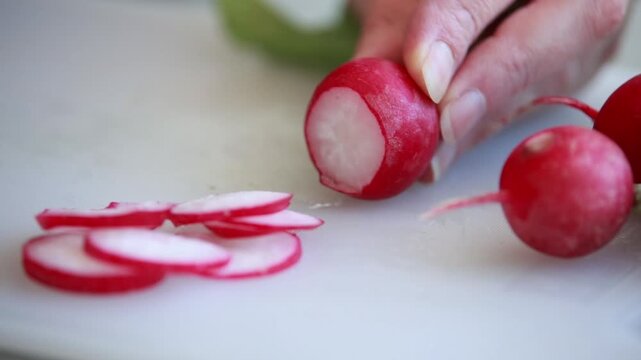 Close-up of a woman's hands slicing fresh red radishes with a chef's knife on a clean white cutting board