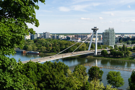 View of the Most SNP bridge with its iconic UFO observation deck crossing the Danube river surrounded by lush greenery and the city skyline Bratislava, Bratislava Region, Slovakia.