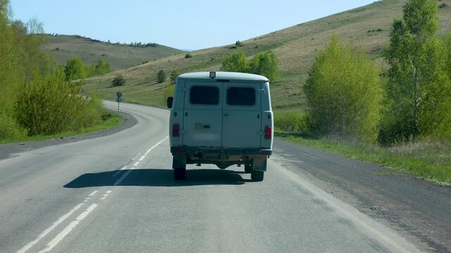 Rear view of a small grey van driving on a paved rural road in a hilly area. 50 FPS 4K footage.