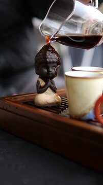 Chinese tea ceremony master pouring hot pu erh tea from a glass pitcher onto a small clay buddha figurine, a traditional ritual for warming the teaware and showing respect before serving