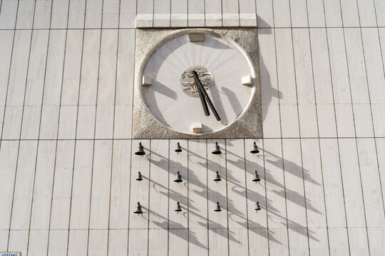 View of a large stone clock and a set of small bells casting shadows on a modern stone facade in Bratislava, Bratislava Region, Slovakia.