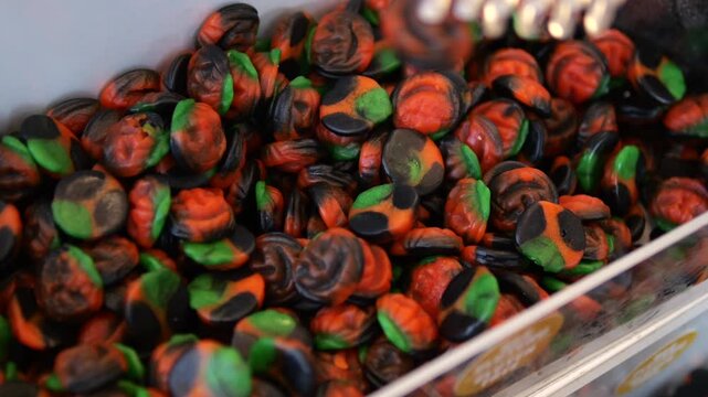 Close up view of a person using plastic tongs to pick up colorful halloween themed gummy candies from a bulk bin in a store, mixing the sugary treats before choosing some