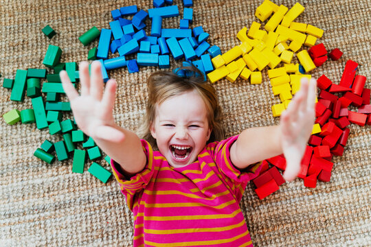 A happy little girl lies down on a carpet surrounded by colorful wooden toy blocks in a playroom. Authentic childhood lifestyle and Montessori heuristic play concept.