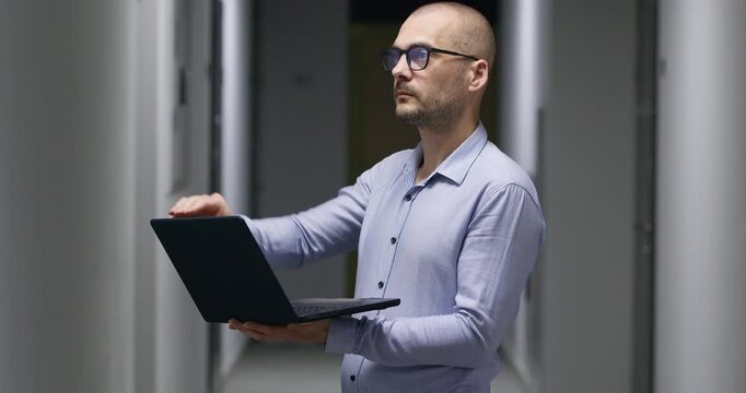 Focused and serious businessman opens sleek laptop in corridor of modern new building. Confident bald man works on important structured document standing in hallway
