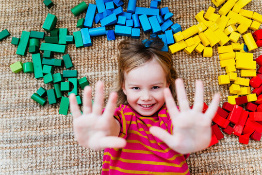 A happy little girl lies down on a carpet surrounded by colorful wooden toy blocks in a playroom. Authentic childhood lifestyle and Montessori heuristic play concept.