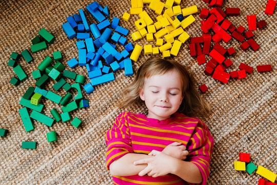A happy little girl lies down on a carpet surrounded by colorful wooden toy blocks in a playroom. Authentic childhood lifestyle and Montessori heuristic play concept.
