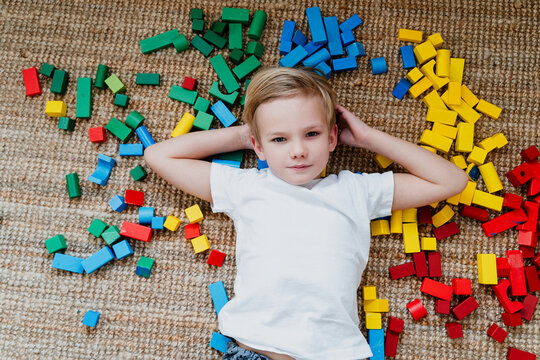 A smiling young boy relaxes on the floor among scattered bright wooden building blocks in a nursery. Authentic domestic life and creative heuristic play development concept.