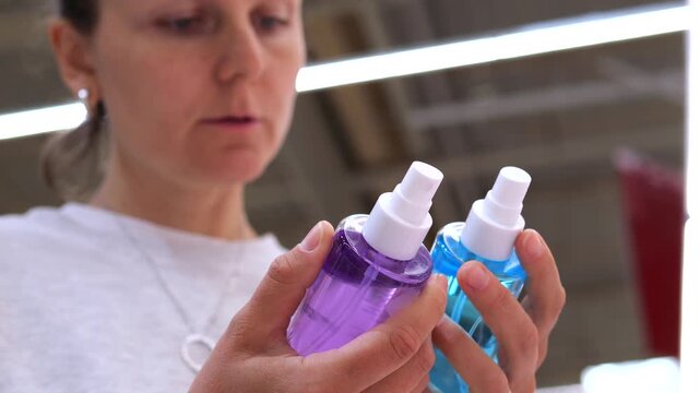 Thoughtful adult woman holding two colorful spray bottles in a store, comparing beauty products and making a difficult choice between blue and purple cosmetic liquid while shopping