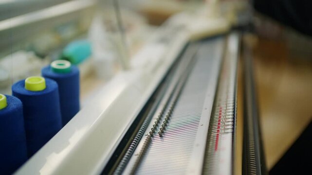 Close up of a knitting machine with metal needles, numbered measuring rail, carriage, and yarn tension system