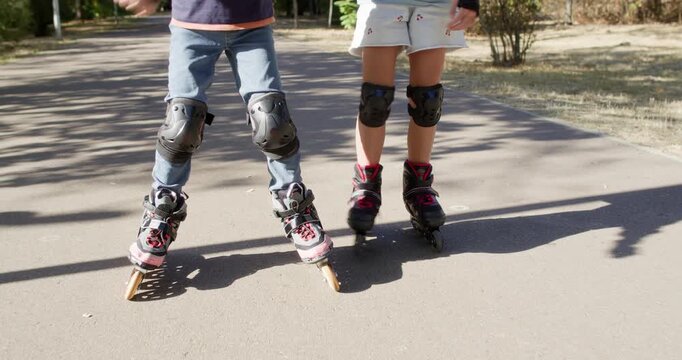 Brother and sister roll slowly on inline rollerblades while holding hands on park path. Children keep knees bent moving carefully to maintain shared balance safely
