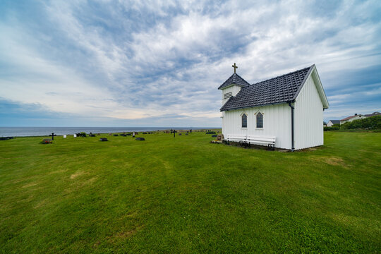 View of the small white Varhaug Old Cemetery chapel standing on a green lawn by the sea under a dramatic cloudy sky in Varhaug, Rogaland, Norway.