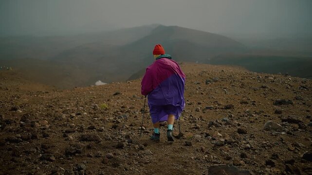 Young man in raincoat and in buff goes with trekking poles along rocky ridge high in mountains. Back view. Slow motion.