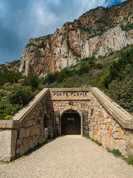 View of the historic stone entrance of Porto Flavia mine nestled against steep rocky cliffs and green vegetation under a blue sky in Iglesias, Sardegna, Italy.