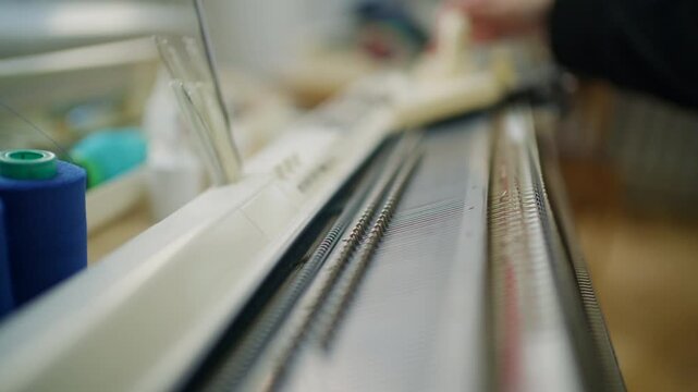 Close up of a knitting machine with metal needles, numbered measuring rail, carriage, and yarn tension system