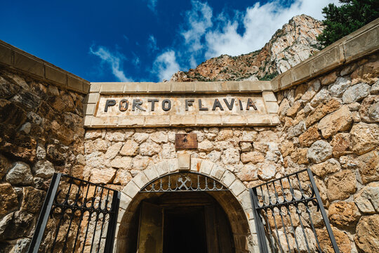 View of the historic entrance of Porto Flavia mining site with its arched stone doorway and rugged mountain backdrop under a blue sky Iglesias, Sardegna, Italy.