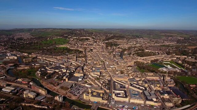 4K cinematic aerial view of Bath with honey-coloured Georgian terraces, Royal Crescent, Bath Abbey, River Avon and surrounding Somerset countryside under clear blue sky.