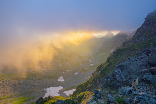 View of Smutne sedlo in Western Tatras with golden sunset light piercing through dense fog over rocky slopes and patches of snow in Ziar, Zilina Region, Slovakia.