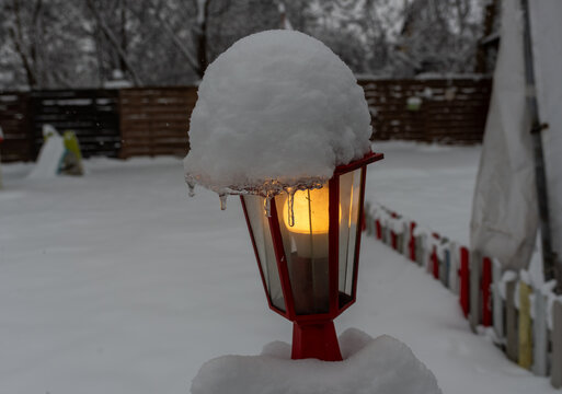 Red outdoor garden lantern glows under a thick cap of fresh snow and icicles, creating a winter patio scene with soft background, cold weather, and warm decorative light.