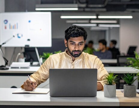 Young Latin professional attending virtual business training on laptop while taking notes and studying in office workspace.