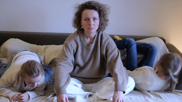 Frustrated mother attempting to practice yoga while her two noisy and unruly children are jumping, playing, and making a mess around her on the sofa, highlighting the challenges of motherhood