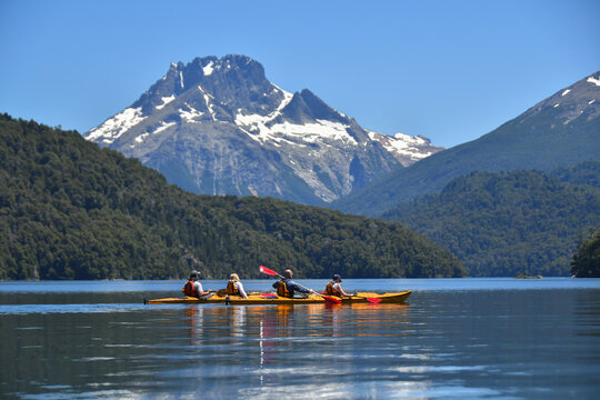 View of people kayaking on the calm waters of Nahuel Huapi Lake with snow-capped mountains and lush forests in San Carlos de Bariloche, Rio Negro, Argentina.