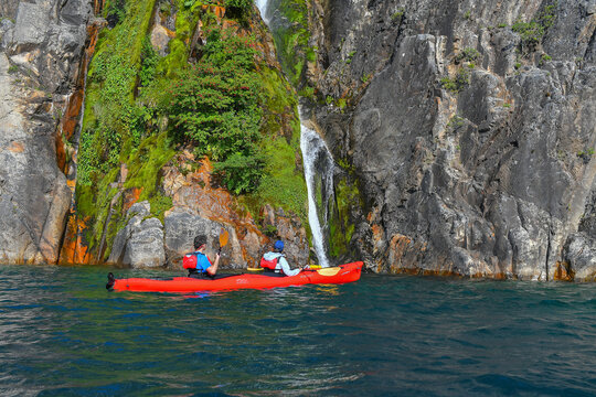 View of two people in a red kayak paddling on the blue waters of Nahuel Huapi Lake near a small waterfall and rocky cliffs San Carlos de Bariloche, Rio Negro, Argentina.