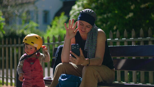 Mother undergoing chemotherapy sitting on park bench waving during video call on smartphone, smiling while toddler hands back tangerine before walking off to play, candid family moment