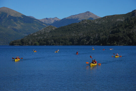 View of people paddling yellow and red kayaks on the blue waters of Nahuel Huapi Lake with forested hills and mountains in San Carlos de Bariloche, Rio Negro, Argentina.