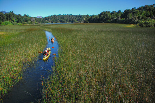 View of people kayaking in a narrow water channel through dense green reeds towards Nahuel Huapi Lake, San Carlos de Bariloche, Rio Negro, Argentina.