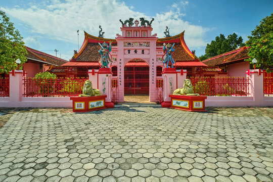 View of Tjoe An Kiong Temple featuring an ornate pink facade, traditional statues, and a stone courtyard under a clear sky in Kecamatan Lasem, Jawa Tengah, Indonesia.