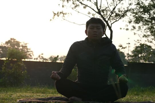 A man sits cross-legged doing meditation and yoga, breathing fresh air, relaxing his body in the park with the silhouette of the soft morning sunlight.