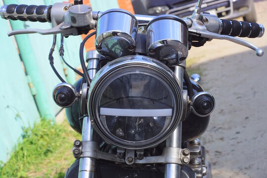 part of an old motorcycle with a white glass headlight and black handlebars near a gas tank on a summer street