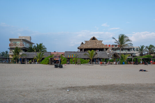 Carrizal, Mexico - December 27 2025: A view of beachfront buildings with thatched roofs and modern structures, surrounded by palm trees, with ATVs on the sandy beach. Guerrero.