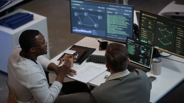 High angle view of diverse male technical specialists analyzing network system while sitting at table with programming codes and diagrams on monitors