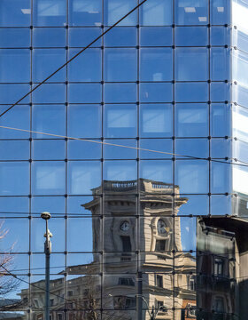 View of the reflection of a historic clock tower building on a modern glass skyscraper facade under a clear blue sky Greater Bilbao, Basque Country, Spain.